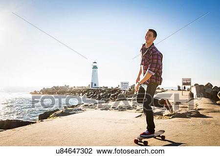 Stock Image - Young man skating on skateboard by water's edge. Fotosearch