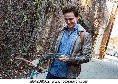 Young man strolling with cycle on sidewalk reading smartphone texts View Large Photo Image Stock Image - Young man strolling with cycle on sidewalk reading smartphone texts. Fotosearch