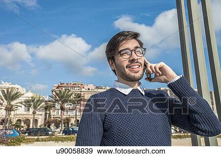 Stock Photo - Young man wearing eyeglasses using smartphone looking away smiling. Fotosearch