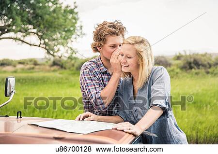 Young man whispering to girlfriend whilst reading map on jeep hood View Large Photo Image Stock Photo - Young man whispering to girlfriend whilst reading map on jeep hood. Fotosearch