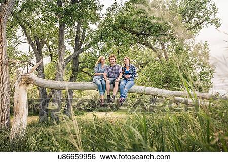 Stock Photograph - Young man with two females sitting together on fallen tree. Fotosearch