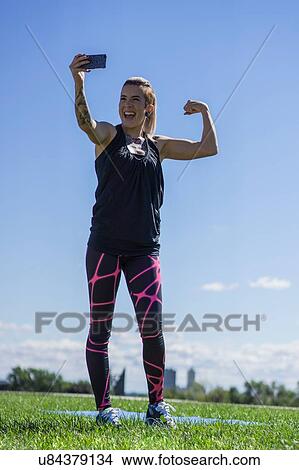 Picture - "Young woman, flexing muscles, taking self portrait, using smartphone, outdoors". Fotosearch