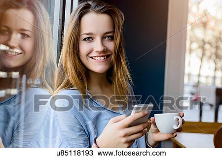 Stock Image - Young woman holding coffee cup and smartphone looking at camera smiling. Fotosearch