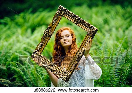 Stock Image - Young woman holding gold picture frame amongst forest ferns. Fotosearch