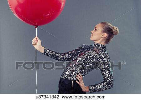 Stock Image - Young woman holding red balloon, grey background. Fotosearch