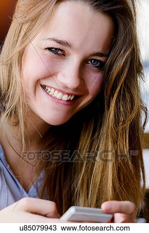 Stock Image - Young woman holding smartphone looking at camera smiling. Fotosearch