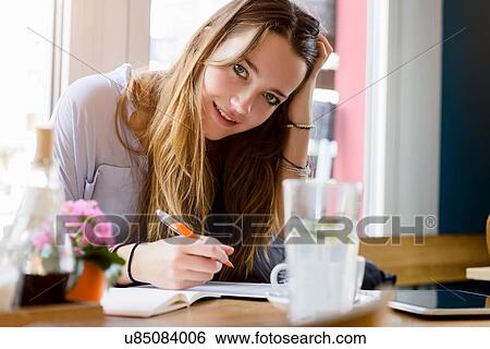 Stock Photograph - Young woman in cafe writing, hand on head resting on elbow looking at camera smiling. Fotosearch