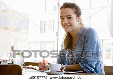 Stock Photography - Young woman in cafe writing, looking at camera smiling. Fotosearch