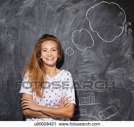 Stock Image - Young woman in front of chalkboard with thought bubble, arms folded looking at camera smiling. Fotosearch