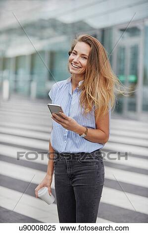 Stock Photography - Young woman in front of pedestrian crossing holding smartphone looking at camera smiling. Fotosearch