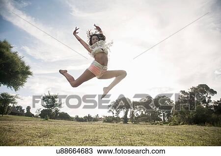 Stock Image - Young woman jumping in mid air, arms raised looking at camera smiling. Fotosearch