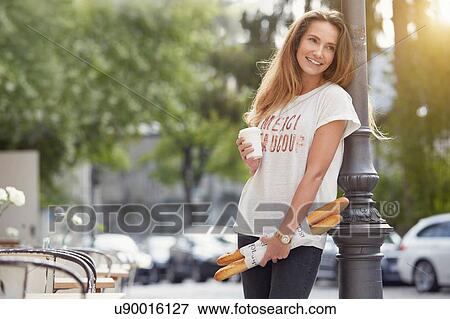 Young woman leaning against lamppost holding baguettes and disposable cup looking away smiling View Large Photo Image Stock Photo - Young woman leaning against lamppost holding baguettes and disposable cup looking away smiling. Fotosearch