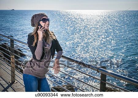 Picture - Young woman leaning against railings by ocean using smartphone looking away smiling. Fotosearch