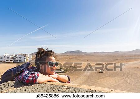 Stock Photograph - Young woman leaning against wall in arid landscape, El Cotillo, Fuerteventura, Spain. Fotosearch