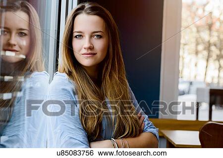 Stock Photograph - Young woman leaning against window arms folded looking at camera smiling. Fotosearch