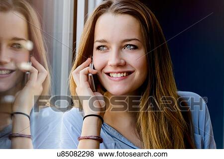 Young woman leaning against window making telephone call looking away smiling View Large Photo Image Stock Photo - Young woman leaning against window making telephone call looking away smiling. Fotosearch