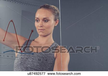 Stock Photo - Young woman looking at chart on glass wall, grey background. Fotosearch