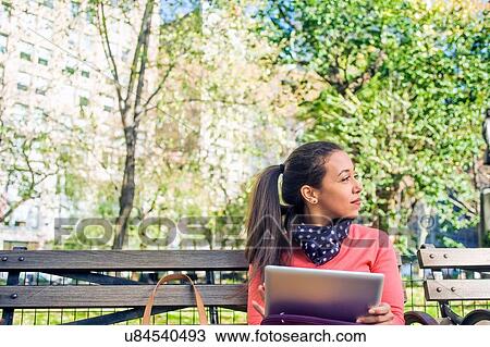Young woman looking sideways from park bench whilst using digital tablet View Large Photo Image Stock Image - Young woman looking sideways from park bench whilst using digital tablet. Fotosearch