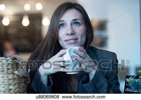 Stock Photo - Young woman looking up whilst drinking coffee in cafe. Fotosearch