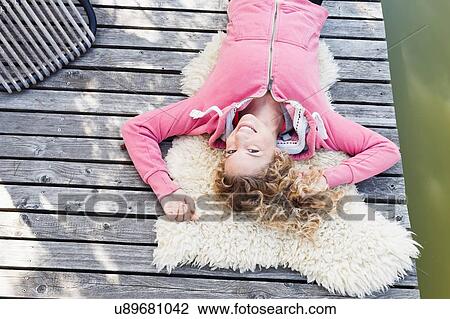 Stock Image - Young woman lying on furry rug on decking, high angle. Fotosearch