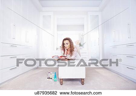 Stock Image - Young woman lying on ottoman reading a book in bedroom. Fotosearch