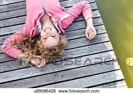 Stock Photograph - Young woman lying on wooden decking smiling to camera. Fotosearch