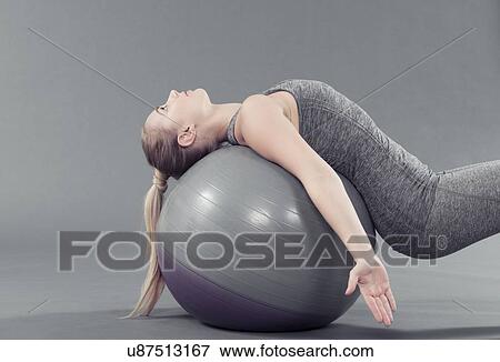 Stock Photo - Young woman practising yoga with gym ball, grey background. Fotosearch