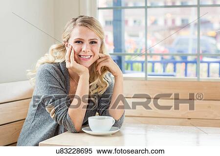 Stock Photography - Young woman sitting at table hand on chin looking at camera smiling. Fotosearch