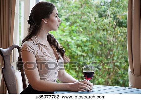 Stock Image - Young woman sitting at table in front of window holding glass of wine looking away. Fotosearch
