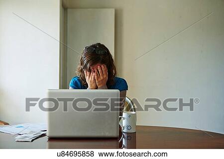 Stock Photo - "Young woman sitting at table using laptop, looking stressed". Fotosearch