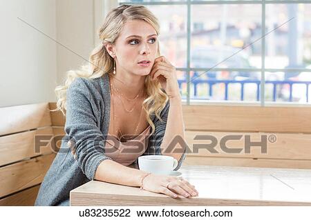 Stock Image - Young woman sitting at table with teacup looking away. Fotosearch