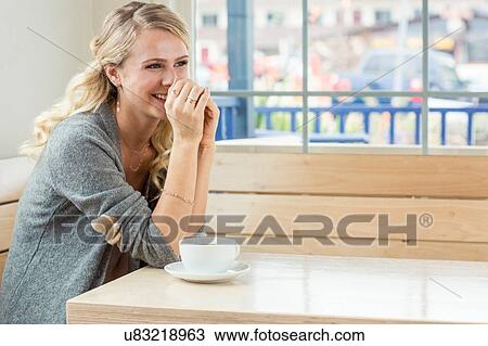 Stock Image - "Young woman sitting at table with tea and saucer, resting on elbows, looking away smiling". Fotosearch
