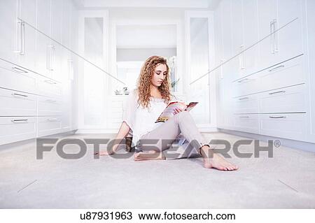 Stock Image - Young woman sitting on floor reading a book in bedroom. Fotosearch