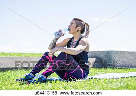 Stock Photo - "Young woman sitting on grass, drinking from water bottle". Fotosearch