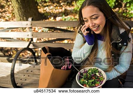 Picture - Young woman sitting on park bench chatting on smartphone whilst eating salad lunch. Fotosearch