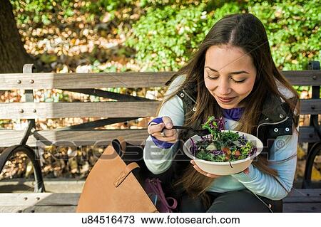 Stock Image - Young woman sitting on park bench eating salad lunch. Fotosearch