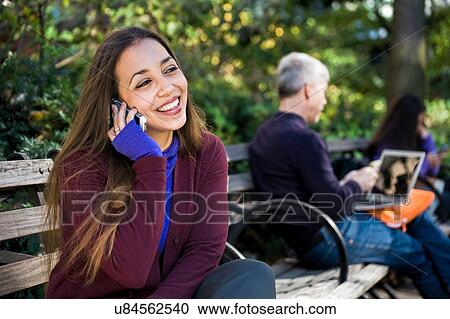 Stock Image - Young woman sitting on park bench chatting on smartphone. Fotosearch