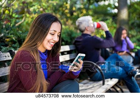 Young woman sitting on park bench reading smartphone texts View Large Photo Image Stock Photo - Young woman sitting on park bench reading smartphone texts. Fotosearch
