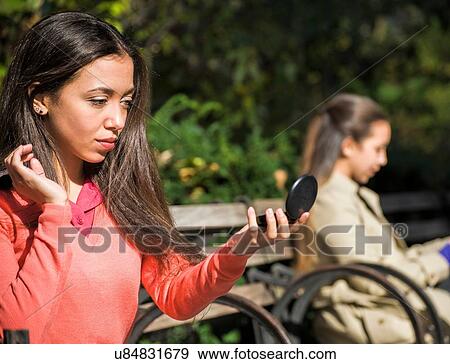 Young woman sitting on park bench looking at compact mirror View Large Photo Image Stock Photo - Young woman sitting on park bench looking at compact mirror. Fotosearch