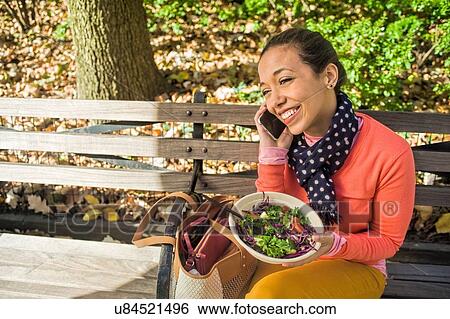 Young woman sitting on park bench chatting on smartphone whilst eating lunch View Large Photo Image Stock Photograph - Young woman sitting on park bench chatting on smartphone whilst eating lunch. Fotosearch