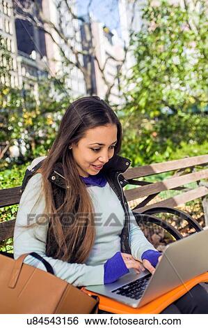 Young woman sitting on park bench typing on laptop View Large Photo Image Stock Photograph - Young woman sitting on park bench typing on laptop. Fotosearch