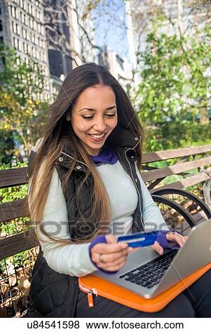 Young woman sitting on park bench using credit card with laptop View Large Photo Image Stock Photo - Young woman sitting on park bench using credit card with laptop. Fotosearch