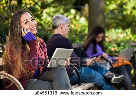 Young woman sitting on park bench using digital tablet and chatting on smartphone View Large Photo Image Stock Photo - Young woman sitting on park bench using digital tablet and chatting on smartphone. Fotosearch