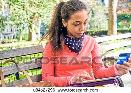 Young woman sitting on park bench using credit card with digital tablet View Large Photo Image Stock Photograph - Young woman sitting on park bench using credit card with digital tablet. Fotosearch
