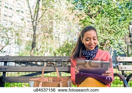 Stock Photograph - Young woman sitting on park bench using digital tablet. Fotosearch