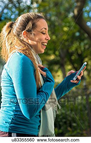 Stock Image - Young woman smiling at message on smartphone in park. Fotosearch