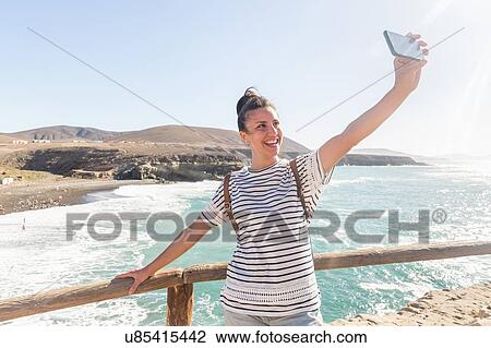 Stock Image - Young woman Taking smartphone selfie with sea, Ajuy, Fuerteventura, Spain. Fotosearch