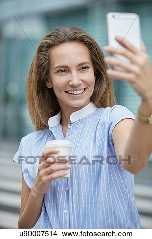 Picture - Young woman using smartphone to take selfie smiling. Fotosearch