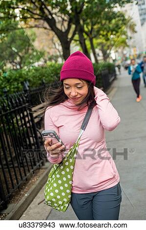 Stock Image - Young woman walking and using smartphone in park. Fotosearch