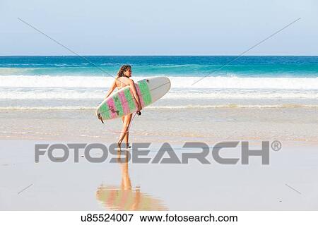 Stock Photo - Young woman walking down to sea carrying surfboard, El Cotillo, Fuerteventura, Spain. Fotosearch